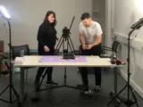 Founders Darren and Lydia stand behind a large desk , making a stop motion animation with a DSLR camera on a tripod. A professional light stands at both ends. 
